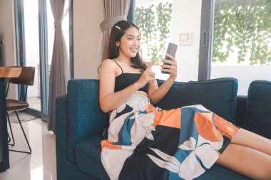 Genç Asyalı kadın boş zamanlarını oturma odasındaki rahat bir koltukta oturarak akıllı telefonundaki mobil uygulamadan mesaj göndererek geçiriyor..