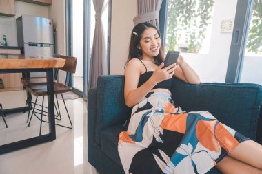 Genç Asyalı kadın boş zamanlarını oturma odasındaki rahat bir koltukta oturarak akıllı telefonundaki mobil uygulamadan mesaj göndererek geçiriyor..