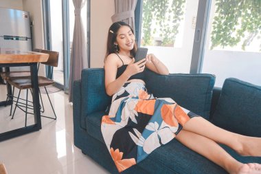 Genç Asyalı kadın boş zamanlarını oturma odasındaki rahat bir koltukta oturarak akıllı telefonundaki mobil uygulamadan mesaj göndererek geçiriyor..