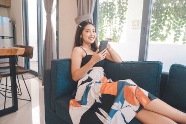 Genç Asyalı kadın boş zamanlarını oturma odasındaki rahat bir koltukta oturarak akıllı telefonundaki mobil uygulamadan mesaj göndererek geçiriyor..