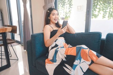 Genç Asyalı kadın boş zamanlarını oturma odasındaki rahat bir koltukta oturarak akıllı telefonundaki mobil uygulamadan mesaj göndererek geçiriyor..