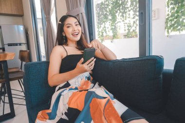 Genç Asyalı kadın boş zamanlarını oturma odasındaki rahat bir koltukta oturarak akıllı telefonundaki mobil uygulamadan mesaj göndererek geçiriyor..