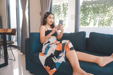 Genç Asyalı kadın boş zamanlarını oturma odasındaki rahat bir koltukta oturarak akıllı telefonundaki mobil uygulamadan mesaj göndererek geçiriyor..