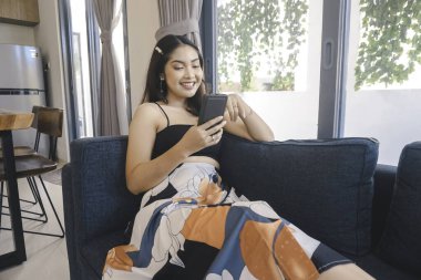 Genç Asyalı kadın boş zamanlarını oturma odasındaki rahat bir koltukta oturarak akıllı telefonundaki mobil uygulamadan mesaj göndererek geçiriyor..