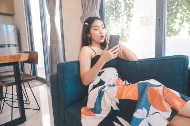 Genç Asyalı kadın boş zamanlarını evde, oturma odasındaki rahat koltukta oturarak geçiriyor. Akıllı telefonuna bakarken şaşırıyor..