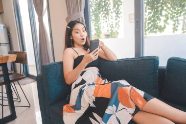 Genç Asyalı kadın boş zamanlarını evde, oturma odasındaki rahat koltukta oturarak geçiriyor. Akıllı telefonuna bakarken şaşırıyor..