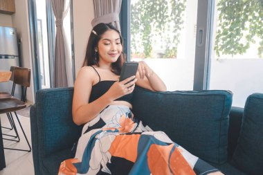 Genç Asyalı kadın boş zamanlarını oturma odasındaki rahat bir koltukta oturarak akıllı telefonundaki mobil uygulamadan mesaj göndererek geçiriyor..
