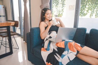 Genç Asyalı kadın zamanını evde, oturma odasındaki rahat bir koltukta oturup kahve içerek ve bilgisayarında online konferans düzenleyerek geçiriyor..