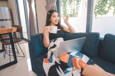 Genç Asyalı kadın zamanını evde, oturma odasındaki rahat bir koltukta oturup kahve içerek ve bilgisayarında online konferans düzenleyerek geçiriyor..