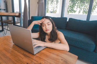 Genç Asyalı kadın zamanını evde oturup hayal kırıklığına uğramış ve yorgun bir şekilde dizüstü bilgisayarı üzerinde çalışarak geçiriyor..