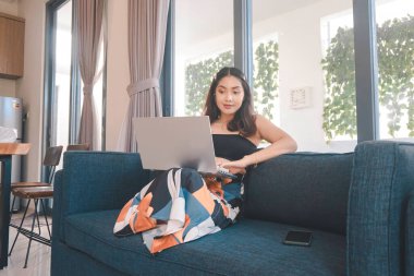 Genç Asyalı kadın zamanını evde oturup gülümseyerek ve dizüstü bilgisayarıyla çalışarak geçiriyor..