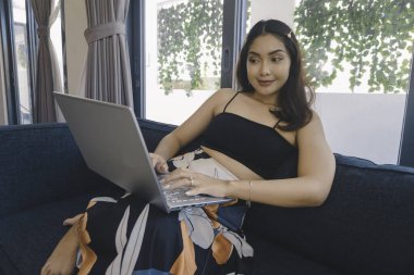 Genç Asyalı kadın zamanını evde oturup gülümseyerek ve dizüstü bilgisayarıyla çalışarak geçiriyor..