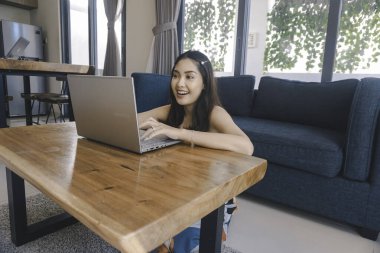 Genç Asyalı kadın zamanını evde oturup gülümseyerek ve dizüstü bilgisayarıyla çalışarak geçiriyor..