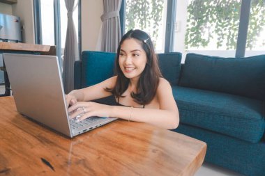 Genç Asyalı kadın zamanını evde oturup gülümseyerek ve dizüstü bilgisayarıyla çalışarak geçiriyor..