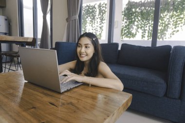 Genç Asyalı kadın zamanını evde oturup gülümseyerek ve dizüstü bilgisayarıyla çalışarak geçiriyor..