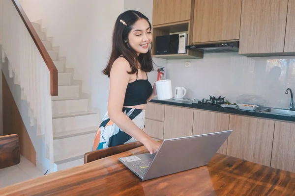 Genç Asyalı kadın zamanını yemek odasında uzaktan çalışarak bilgisayarında kupa taşırken çevrimiçi konferans yaparak geçiriyor..