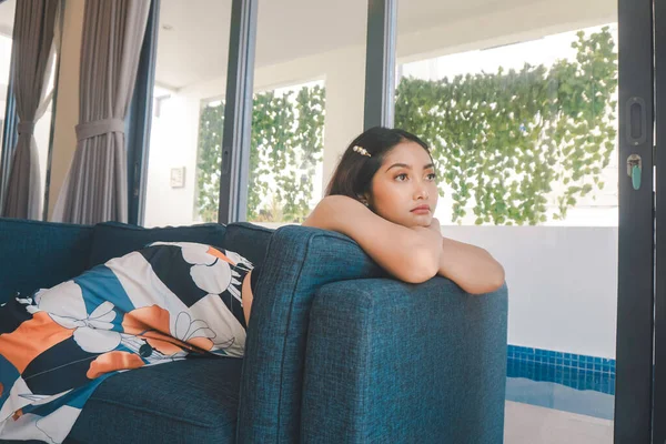 Young Asian woman sitting on the couch in the living room looks sorrowful.