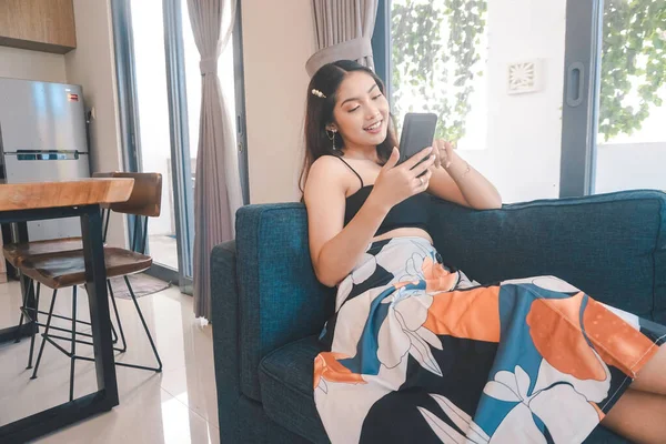 Genç Asyalı kadın boş zamanlarını oturma odasındaki rahat bir koltukta oturarak akıllı telefonundaki mobil uygulamadan mesaj göndererek geçiriyor..