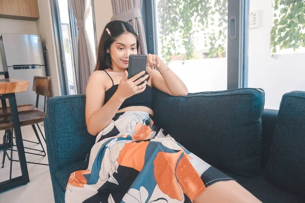 Genç Asyalı kadın boş zamanlarını oturma odasındaki rahat bir koltukta oturarak akıllı telefonundaki mobil uygulamadan mesaj göndererek geçiriyor..