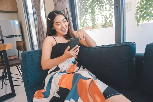 Genç Asyalı kadın boş zamanlarını oturma odasındaki rahat bir koltukta oturarak akıllı telefonundaki mobil uygulamadan mesaj göndererek geçiriyor..