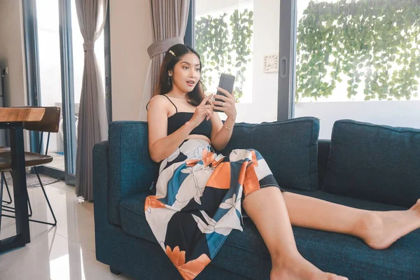 Genç Asyalı kadın boş zamanlarını oturma odasındaki rahat bir koltukta oturarak akıllı telefonundaki mobil uygulamadan mesaj göndererek geçiriyor..