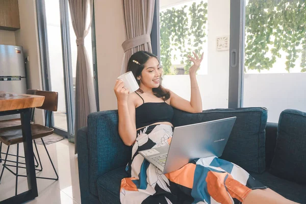 Genç Asyalı kadın zamanını evde, oturma odasındaki rahat bir koltukta oturup kahve içerek ve bilgisayarında online konferans düzenleyerek geçiriyor..