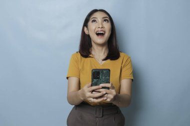 An excited young woman is smiling while holding her smartphone in her hand, isolated on blue background