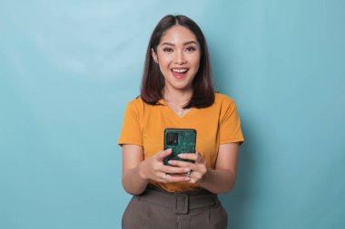 An excited young woman is smiling while holding her smartphone in her hand, isolated on blue background