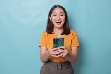 An excited young woman is smiling while holding her smartphone in her hand, isolated on blue background