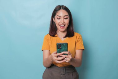 An excited young woman is smiling while holding her smartphone in her hand, isolated on blue background