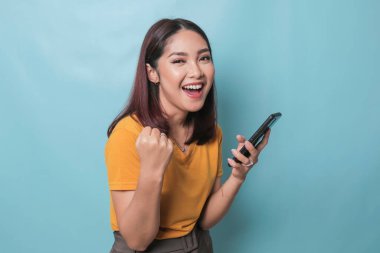 An excited young woman is smiling while holding her smartphone in her hand, isolated on blue background