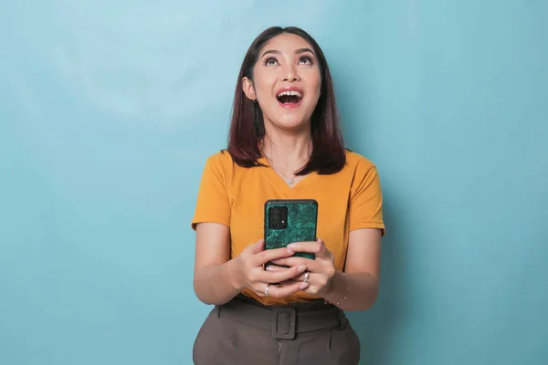 An excited young woman is smiling while holding her smartphone in her hand, isolated on blue background