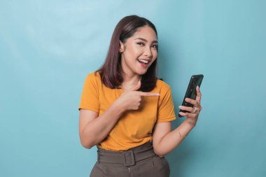 An excited young woman pointing her smartphone in her hand, isolated on blue background