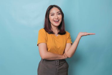 An excited young woman presenting and pointing copy space on her side, isolated on blue background
