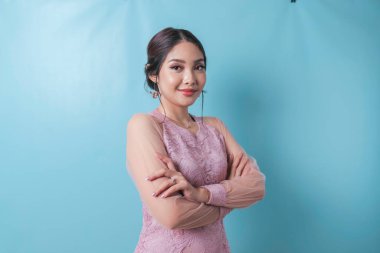 Portrait of a confident smiling girl standing with arms folded and looking at camera isolated over blue background, wearing modern kebaya dress