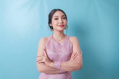 Portrait of a confident smiling girl standing with arms folded and looking at camera isolated over blue background, wearing modern kebaya dress