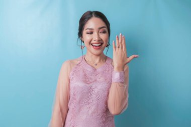 Smiling young Asian woman wearing modern kebaya showing her engagement ring isolated over blue background