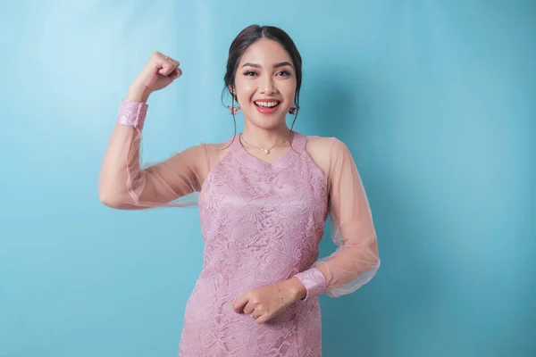 Excited Asian woman wearing modern kebaya clothes showing strong gesture by lifting her arms and muscles smiling proudly