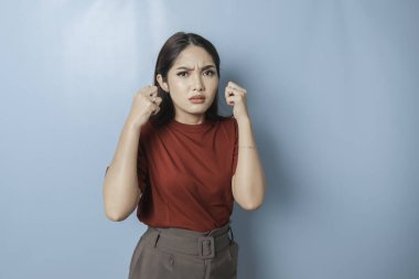 A dissatisfied young Asian woman looks at the camera posing on a blue background, disgruntled girl wearing red t-shirt irritated face expressions show negative attitude concept image