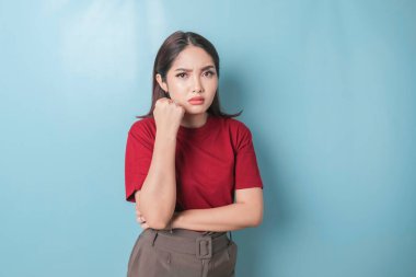 A dissatisfied young Asian woman looks at the camera posing on a blue background, disgruntled girl wearing red t-shirt irritated face expressions show negative attitude concept image