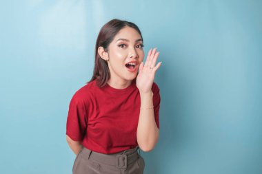 A portrait of an excited Asian woman wearing read t-shirt shouting, isolated by blue background