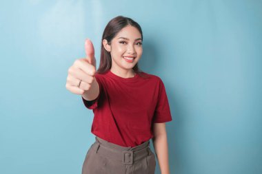 Excited Asian woman wearing a red t-shirt gives thumbs up hand gesture of approval, isolated by blue background