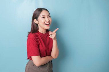 Excited Asian woman wearing red t-shirt pointing at the copy space beside her, isolated by blue background