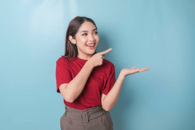 Excited Asian woman wearing red t-shirt pointing at the copy space beside her, isolated by blue background