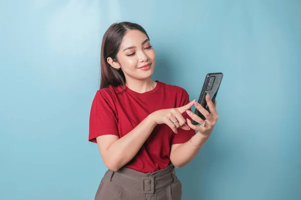 Excited Asian woman wearing a red t-shirt pointing to her smartphone, isolated by a blue background
