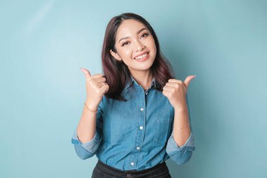 Excited Asian woman wearing a blue shirt gives thumbs up hand gesture of approval, isolated by blue background