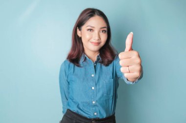 Excited Asian woman wearing a blue shirt gives thumbs up hand gesture of approval, isolated by blue background