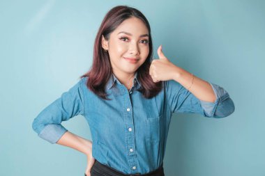Excited Asian woman wearing a blue shirt gives thumbs up hand gesture of approval, isolated by blue background