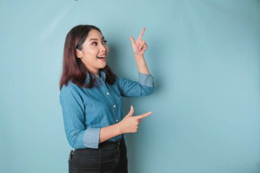 Excited Asian woman wearing blue shirt pointing at the copy space beside her, isolated by blue background