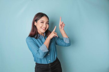 Excited Asian woman wearing blue shirt pointing at the copy space beside her, isolated by blue background